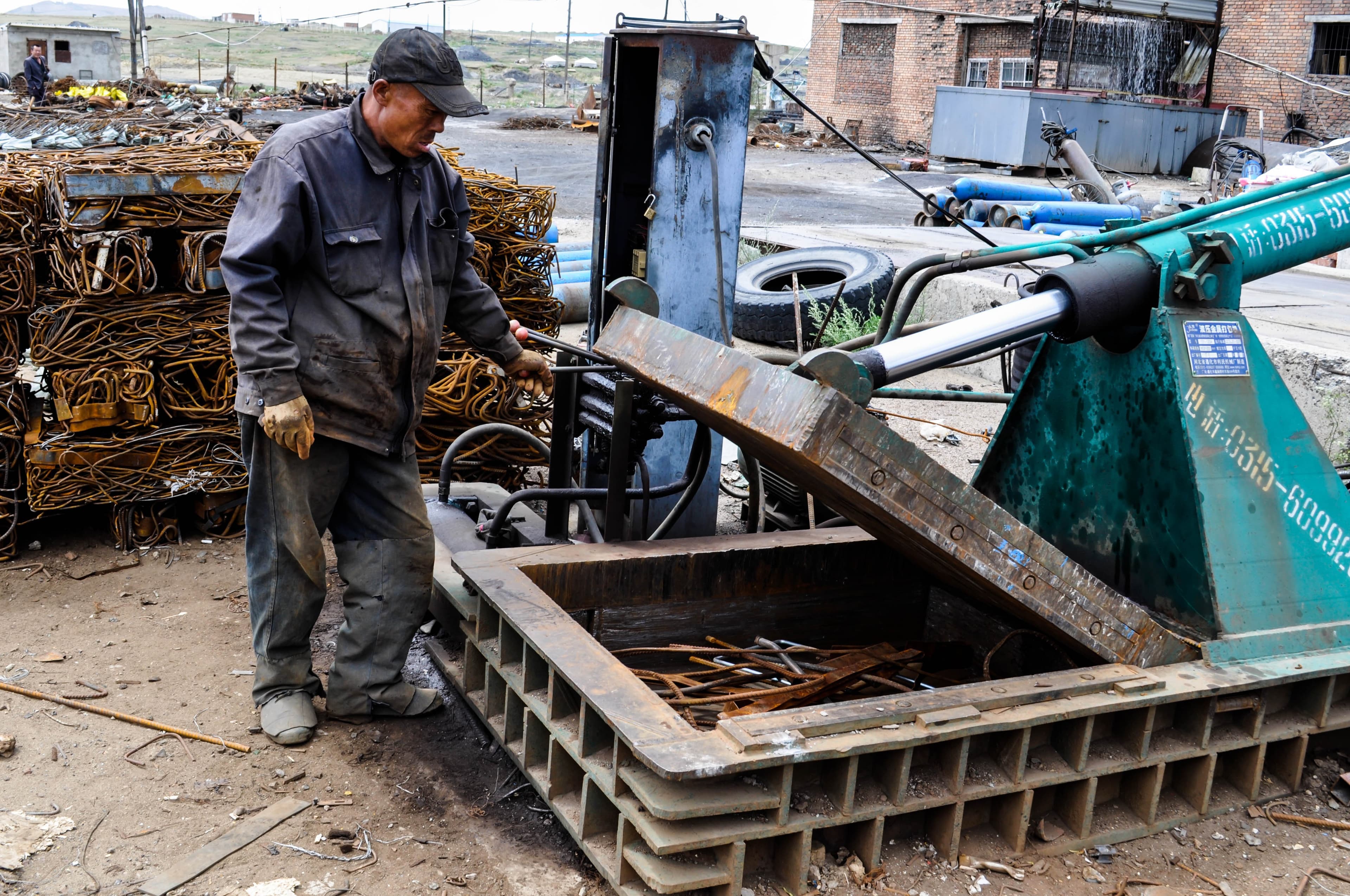A worker at a scrap baling press in a yard stacked with bundled steel.